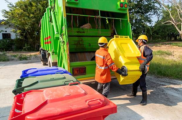 Worker loading household trash bags into a wheeled bin beside driveway